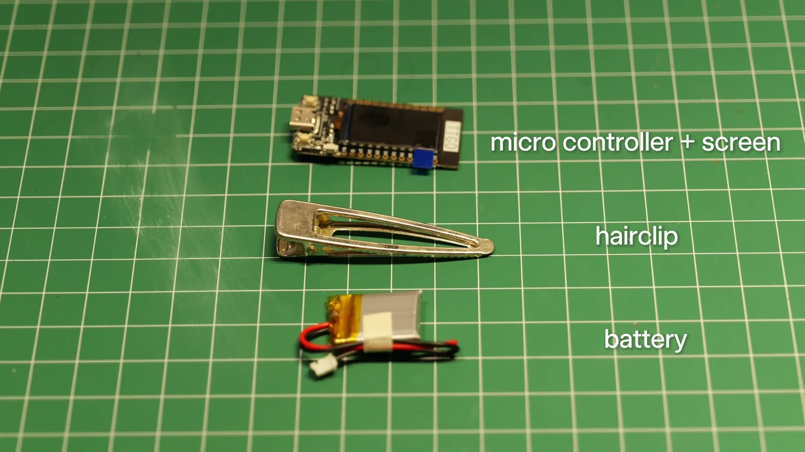 The three parts laid out on a cutting mat: the microcontroller with its screen, a gold metal hair clip, and the LiPo battery.